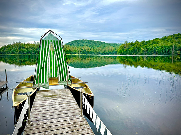 A green-striped tent set up on a wooden platform between two canoes, floating by the dock on the calm Raquette River with forested hills in the background.