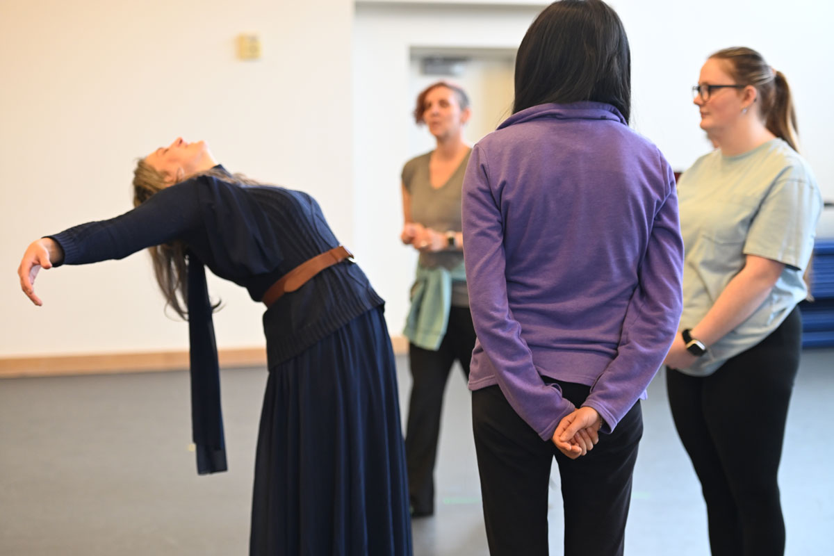 Participants indoors engage in a movement activity, one dancer leaning back dramatically while others observe and prepare to move.