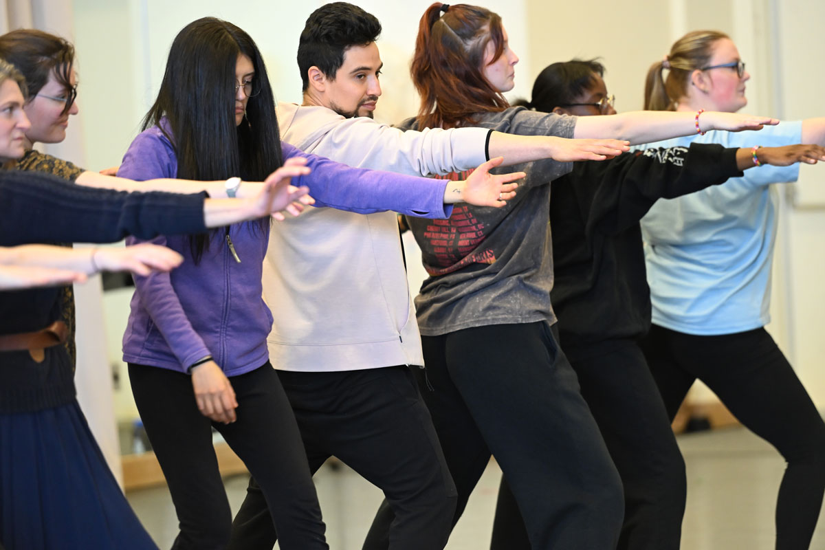 Participants indoors stand in a row with arms extended forward, moving together as part of a dance-like movement choir activity.