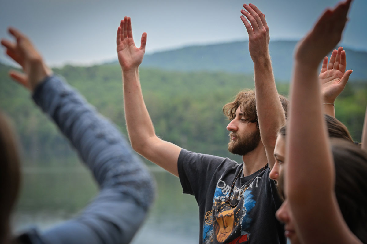 A male participant with raised arms joins others in a movement choir session by the Raquette River, with blurred participants’ arms in the foreground.
