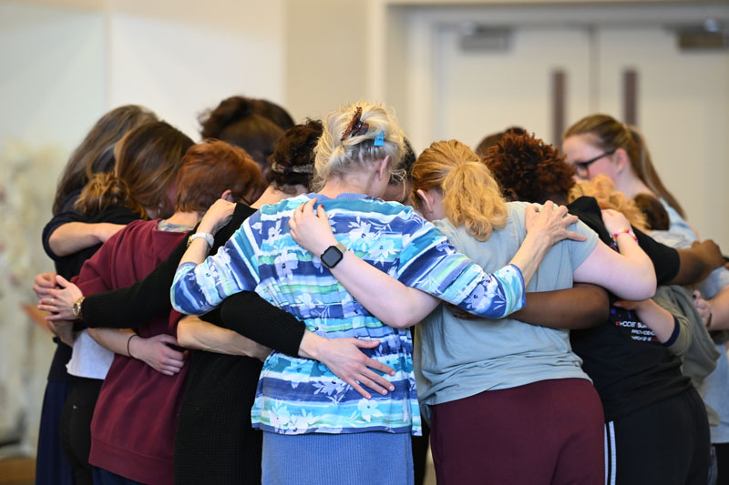 A circle of participants huddles closely together with arms around each other during a movement choir session.